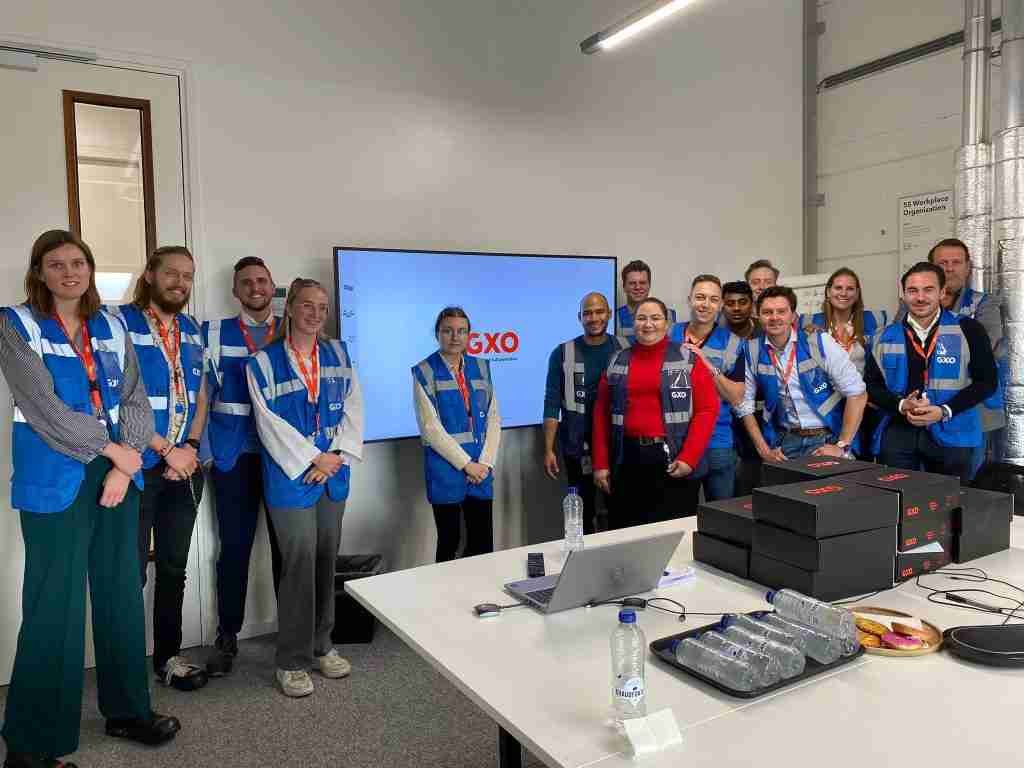 Een groep in blauwe veiligheidsvesten en zakelijke kleding staat voor een scherm met GXO in een vergaderruimte