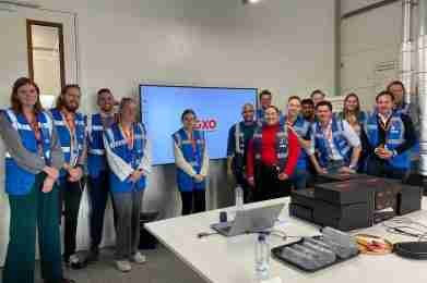 Een groep in blauwe veiligheidsvesten en zakelijke kleding staat voor een scherm met GXO in een vergaderruimte