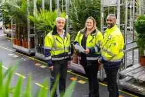 Foto van drie mensen met een gele jas voor een kar met bloemen